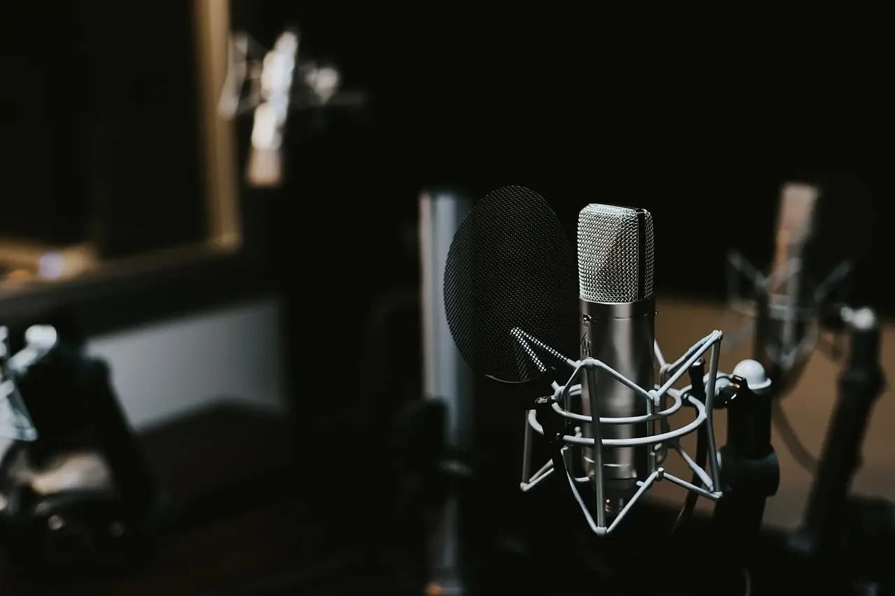 Close-up of a silver microphone with a pop filter in a dimly lit recording studio, surrounded by other audio equipment.